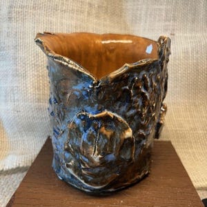 May include: A handcrafted ceramic vase with a textured, organic design. The exterior features a blend of blue, brown, and black hues, while the interior is a warm brown. The vase sits on a brown wooden surface.
