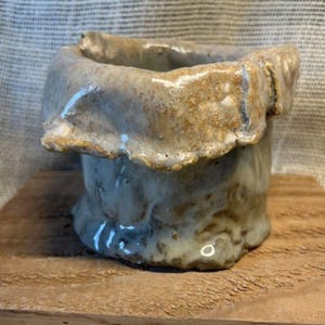 May include: A small, hand-formed ceramic vessel with an irregular, folded rim. The piece features a glossy, mottled glaze in shades of beige, tan, and light gray, with a textured, uneven surface that highlights the organic shape of the clay body.