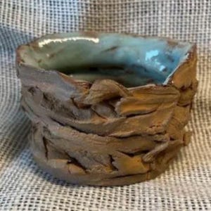 May include: Handmade ceramic bowl with a textured, brown exterior and a turquoise interior. The bowl has a rough, organic shape, suggesting a rustic or artisanal aesthetic. The rim is uneven, adding to its unique character.