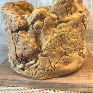 Op de afbeelding: Handgemaakte keramische pot met een getextureerde, aardse uitstraling. De pot is voornamelijk bruin en beige, met een ruw, ongelijkmatig oppervlak. Hij staat op een kleine houten basis, wat een rustieke of natuurlijke uitstraling geeft.