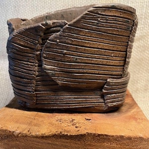 May include: A handcrafted, brown ceramic vessel with a textured, ridged surface. The vessel has a unique, folded design and sits on a wooden base. The earthy tones and organic shape suggest a decorative or functional art piece.