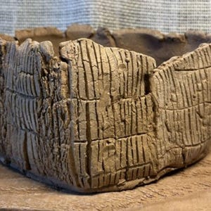 May include: A hand-built, unglazed clay bowl with a rough, textured exterior featuring deep vertical and horizontal grooves. The bowl has an irregular, organic shape with a thick, uneven rim and a natural, earthy brown color throughout the surface.