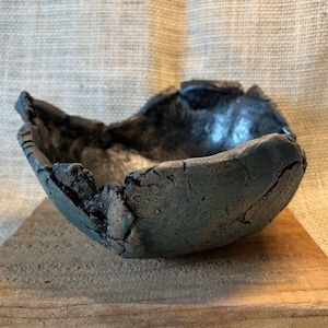 May include: A handcrafted, dark gray ceramic bowl with a rough, textured surface and a cracked design. The bowl is placed on a wooden surface, showcasing its unique, organic shape. The interior has a smooth, reflective surface.
