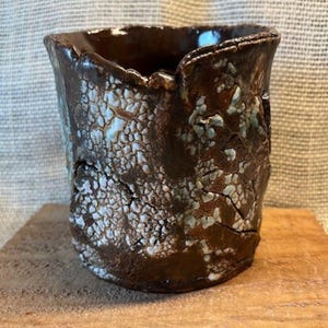 May include: A handmade ceramic mug with a textured surface. The mug is dark brown with white and light blue accents, and a cracked pattern. The rim of the mug has a unique, uneven shape. The mug is sitting on a wooden surface.