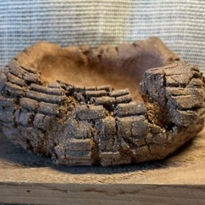 May include: A small, round, hand-formed ceramic bowl with a dark brown, textured exterior that mimics the appearance of rough tree bark. The interior of the bowl is smooth and concave, featuring a lighter brown, matte clay finish throughout.