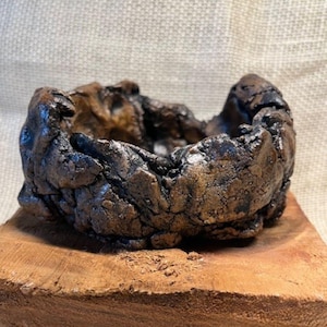 May include: A handcrafted, small, brown and black ceramic bowl with a rough, textured surface. The bowl is set on a wooden base, showcasing its organic shape and earthy tones. The bowl's interior is dark, contrasting with the lighter brown exterior.