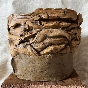 Puede incluir: Una maceta de cerámica hecha a mano con un diseño texturizado y en capas en tonos marrones y beige. La parte superior tiene una apariencia rugosa y orgánica, mientras que la base es una banda lisa y moteada. La maceta se encuentra sobre un pequeño bloque de madera marrón rojizo.