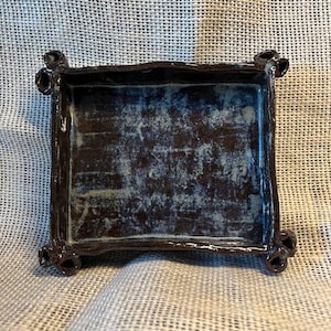 May include: A square, handmade ceramic tray with a dark brown exterior and a mottled blue and brown interior. The tray has a raised rim and decorative corner accents. The ceramic piece is displayed on a textured, off-white surface.