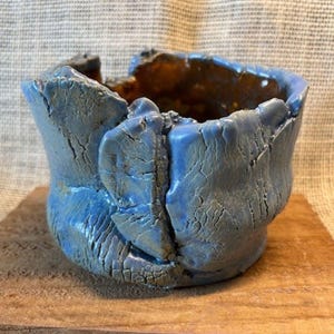 May include: A handmade, blue ceramic pot with a textured surface and a rough, uneven rim. The interior has a contrasting brown color. The pot is displayed on a wooden surface, showcasing its unique, organic form.