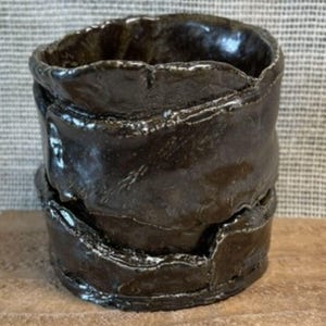May include: A dark brown ceramic cup with a glossy, reflective finish. The vessel features a hand-formed, uneven, and layered texture with prominent ridges and a thick, irregular rim, creating a distinctively organic and sculptural appearance.