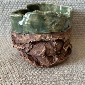 Puede incluir: Un jarrón de cerámica hecho a mano con un diseño orgánico y texturizado. La parte superior está esmaltada en verde brillante, mientras que la sección inferior presenta una textura marrón terrosa rugosa. El jarrón tiene un borde abierto irregular.