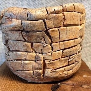 May include: A cylindrical ceramic vessel with a light tan, textured surface featuring deep horizontal grooves and vertical cracks throughout the exterior. The bowl has a thick, uneven rim and a rounded base, displaying a weathered, earthy appearance.