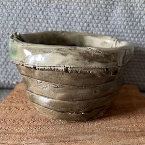 May include: A handmade ceramic bowl with a textured, layered design. The bowl features a muted color palette of olive green and beige, with a slightly irregular rim. The bowl is sitting on a wooden surface.