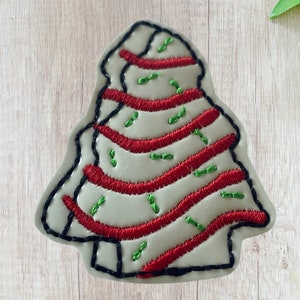 May include: A white felt Christmas tree applique with red and green embroidery. The tree is decorated with red stripes and green sprinkles.