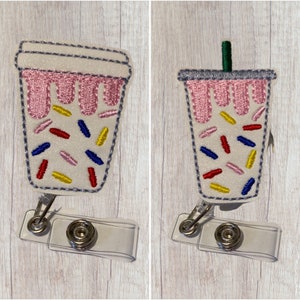 May include: Two white and pink iced coffee cup badge reels with a clear plastic badge holder. The coffee cups have sprinkles and a pink drizzle.