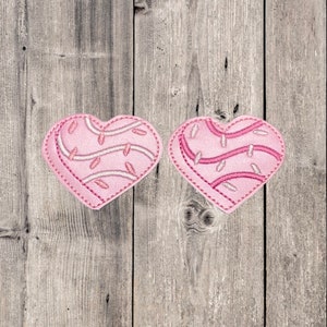 May include: Two pink felt heart-shaped patches with white and pink embroidery. The hearts are on a gray wooden background.