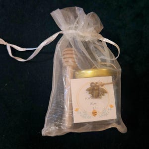 May include: A small jar of honey with a wooden honey dipper inside a sheer white gift bag. The jar has a gold lid and a label with a bee illustration and the words "For Here". The bag is tied with a white ribbon.