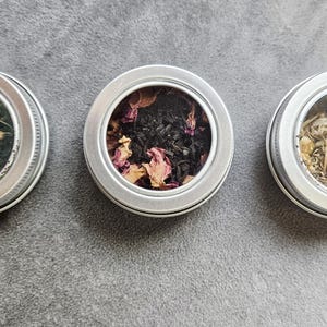 May include: Three small, round, silver tins with clear lids, each filled with different dried herbs and flowers. The contents include green, black, and beige mixtures, suggesting a variety of teas or herbal blends. The tins are arranged in a row on a gray surface.