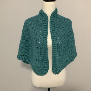 Puede incluir: Un chal de ganchillo de color turquesa, exhibido en un maniquí. El chal de frente abierto presenta un patrón de puntadas texturizadas y una forma redondeada. El escote es un cuello doblado. El chal está hecho de un hilo suave.