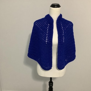 Puede incluir: Un chal de ganchillo azul real con frente abierto, exhibido en un maniquí. El chal presenta un patrón de puntadas texturizadas y un cuello. La prenda está hecha de un hilo suave y es adecuada para usar en capas.
