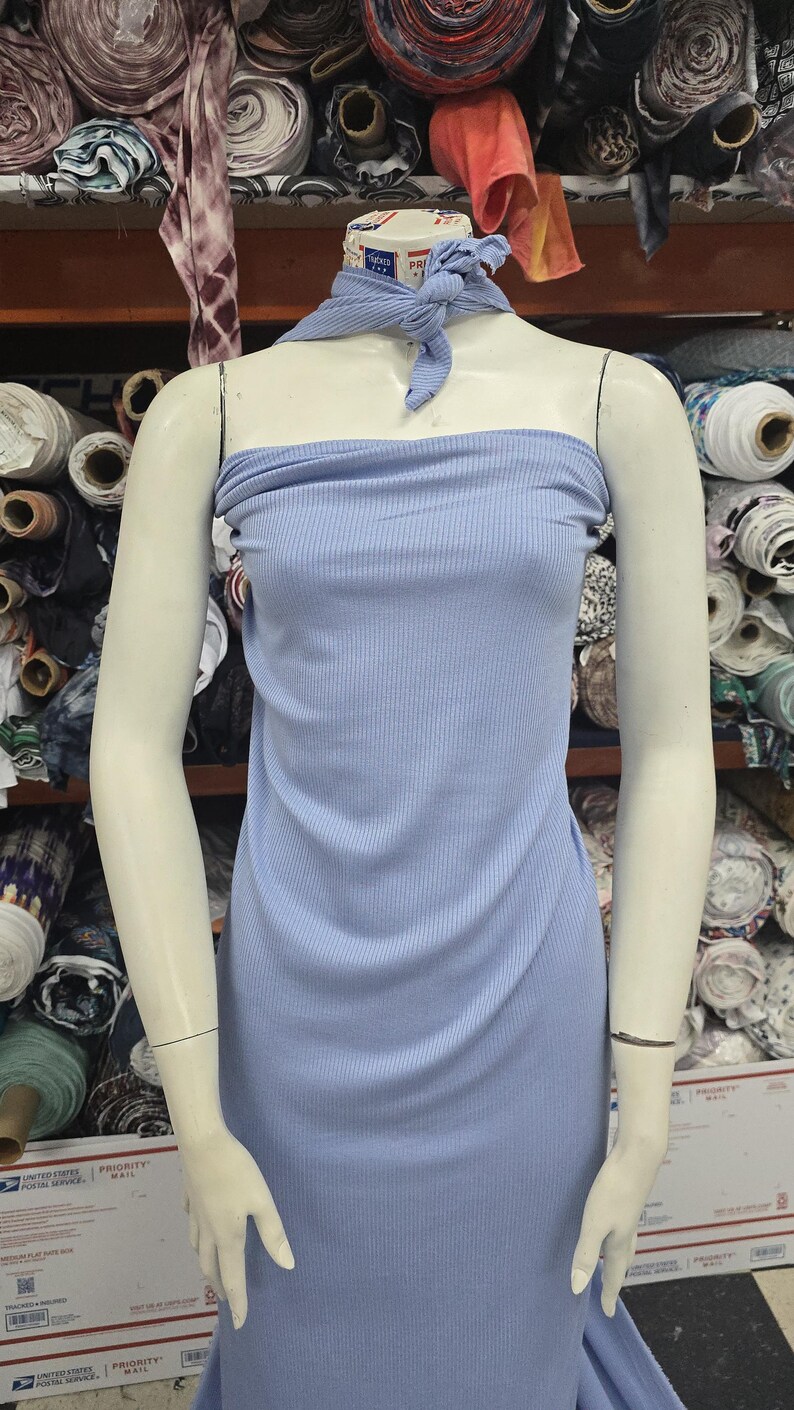 Puede incluir: Un vestido tubo sin tirantes de color azul claro, confeccionado en tejido acanalado el&aacute;stico. La prenda incluye un pa&ntilde;uelo de cuello a juego de tejido acanalado azul claro anudado en la parte delantera, creando un conjunto coordinado en un maniqu&iacute;.