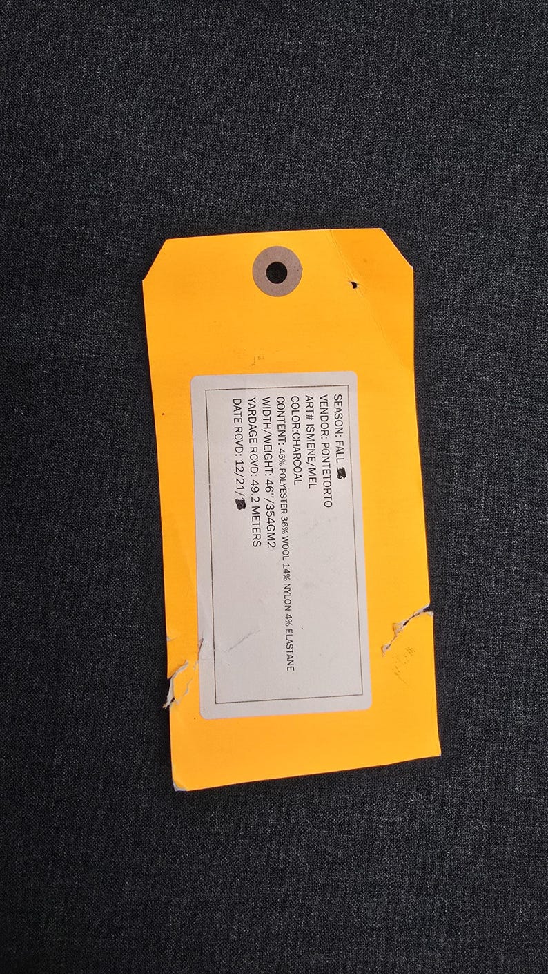 May include: An orange tag with a hole at the top, attached to a dark gray fabric. The tag contains white text with details about the fabric's composition, including 46% polyester, 36% wool, and 14% nylon. The fabric width is 46 inches.