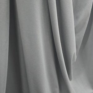 May include: A solid light gray fabric featuring a smooth, matte texture with deep, vertical folds and soft shadows that create a draped effect across the entire surface, highlighting the consistent color and uniform weave of the material.