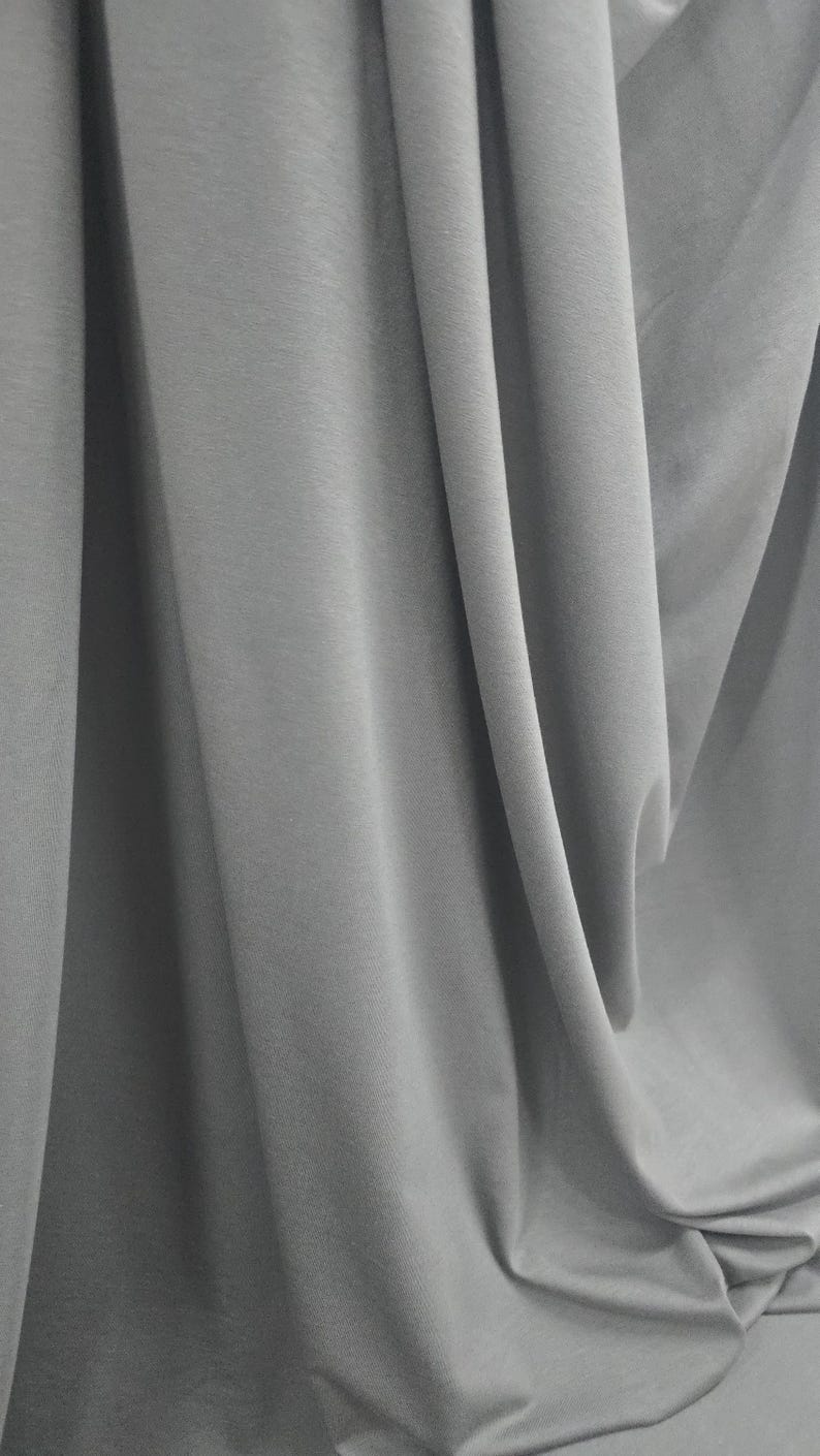 May include: A solid light gray fabric featuring a smooth, matte texture with deep, vertical folds and soft shadows that create a draped effect across the entire surface, highlighting the consistent color and uniform weave of the material.