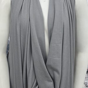 May include: A solid grey fabric draped over a white mannequin. The fabric features a soft, matte texture and hangs in loose, vertical folds from the neck area down the torso, creating a layered appearance with a deep, central cowl neckline.