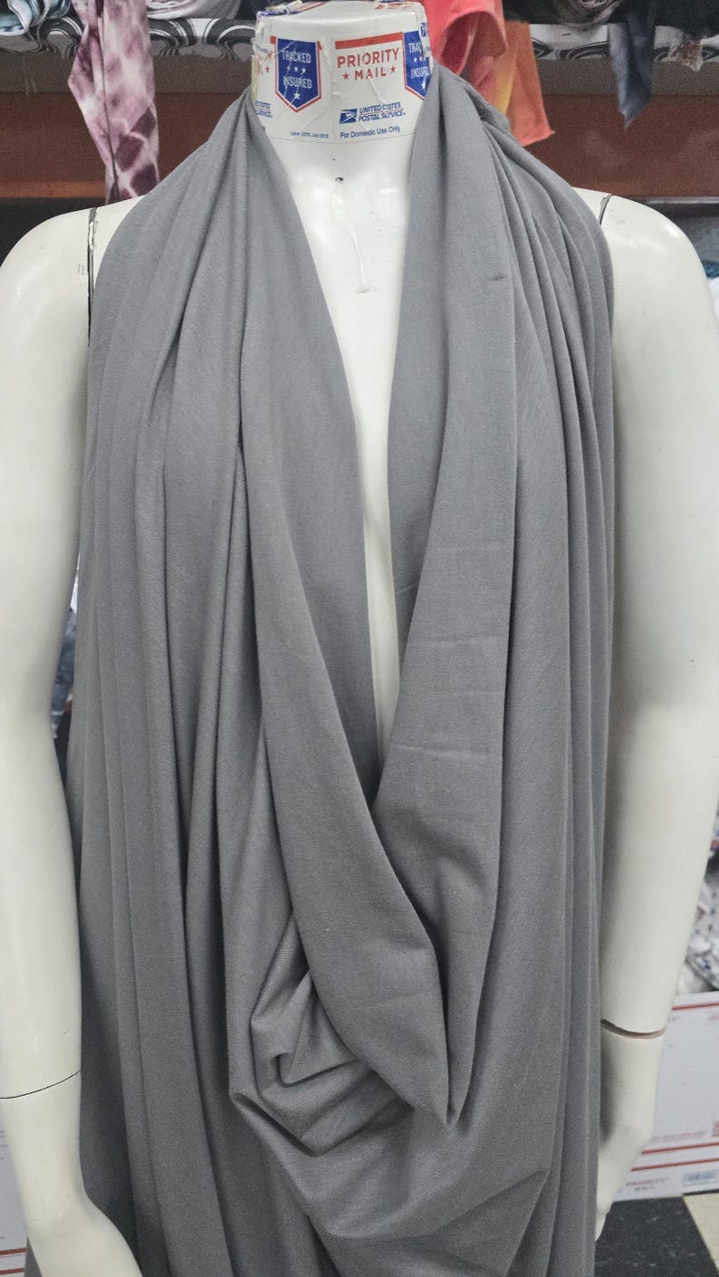 May include: A solid grey fabric draped over a white mannequin. The fabric features a soft, matte texture and hangs in loose, vertical folds from the neck area down the torso, creating a layered appearance with a deep, central cowl neckline.