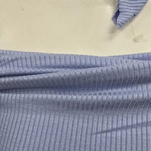 Puede incluir: Una prenda superior de punto acanalado en color azul claro con escote descubierto y una bufanda o lazo de tela acanalada a juego en azul claro drapeado alrededor de la parte superior, mostrando una textura vertical consistente en toda la pieza.