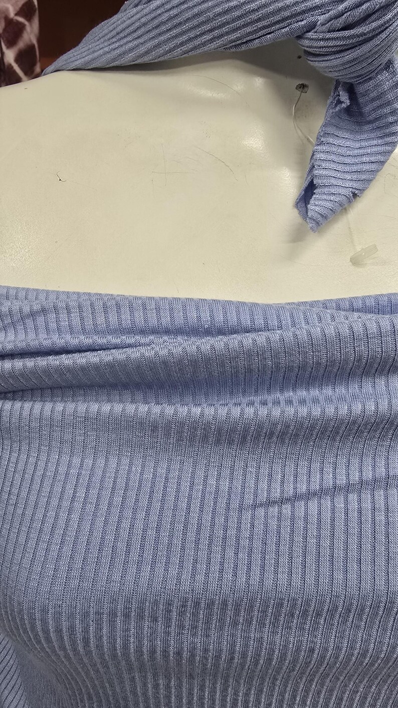 Puede incluir: Una prenda superior de punto acanalado en color azul claro con escote descubierto y una bufanda o lazo de tela acanalada a juego en azul claro drapeado alrededor de la parte superior, mostrando una textura vertical consistente en toda la pieza.