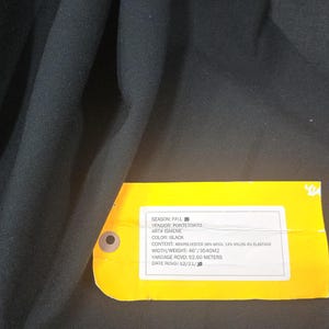 May include: Black fabric with a yellow tag attached. The tag reads "SEASON: FALL, COLOR: BLACK, CONTENT: 46% POLYESTER 36% WOOL 14% NYLON 4% ELASTANE, YARDAGE RCVD: 52.60 METERS".