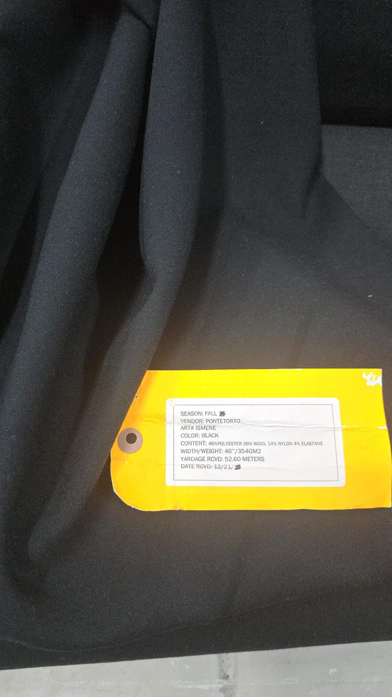 May include: Black fabric with a yellow tag attached. The tag reads "SEASON: FALL, COLOR: BLACK, CONTENT: 46% POLYESTER 36% WOOL 14% NYLON 4% ELASTANE, YARDAGE RCVD: 52.60 METERS".
