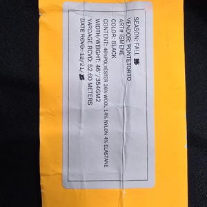 May include: A yellow tag with a hole at the top, featuring text about fabric content and specifications. The tag includes details such as the vendor, color (black), and material composition: 46% polyester, 36% wool, 14% nylon, and 4% elastane.