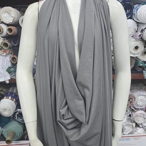May include: A solid grey fabric draped over a mannequin. The fabric features a deep cowl neckline that creates soft, vertical folds extending down the front. The material appears smooth and matte, covering the torso and shoulders completely.