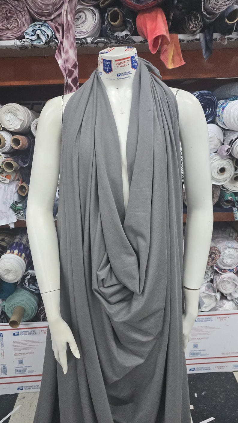 May include: A solid grey fabric draped over a mannequin. The fabric features a deep cowl neckline that creates soft, vertical folds extending down the front. The material appears smooth and matte, covering the torso and shoulders completely.