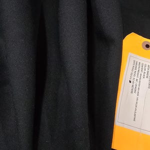 May include: Black fabric with a tag attached. The tag provides details about the fabric's composition, including 46% polyester, 36% wool, 14% nylon, and 4% elastane. The fabric width is 46 inches, and the weight is 354 grams per square meter.