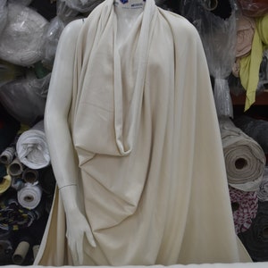 May include: A cream-colored fabric draped over a mannequin. The fabric is soft and flowing, with a subtle sheen. The fabric is displayed against a background of various rolls of fabric in different colors and patterns.