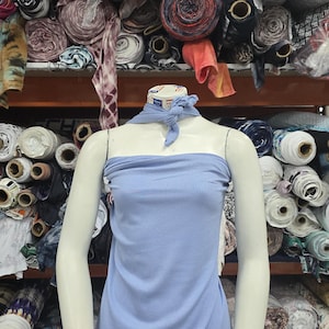 Puede incluir: Un vestido tubo de color azul claro acanalado sobre un maniqu&iacute;, con un pa&ntilde;uelo de cuello a juego de tela acanalada azul claro anudado en la parte delantera. El maniqu&iacute; est&aacute; colocado frente a estantes apilados con rollos de telas.
