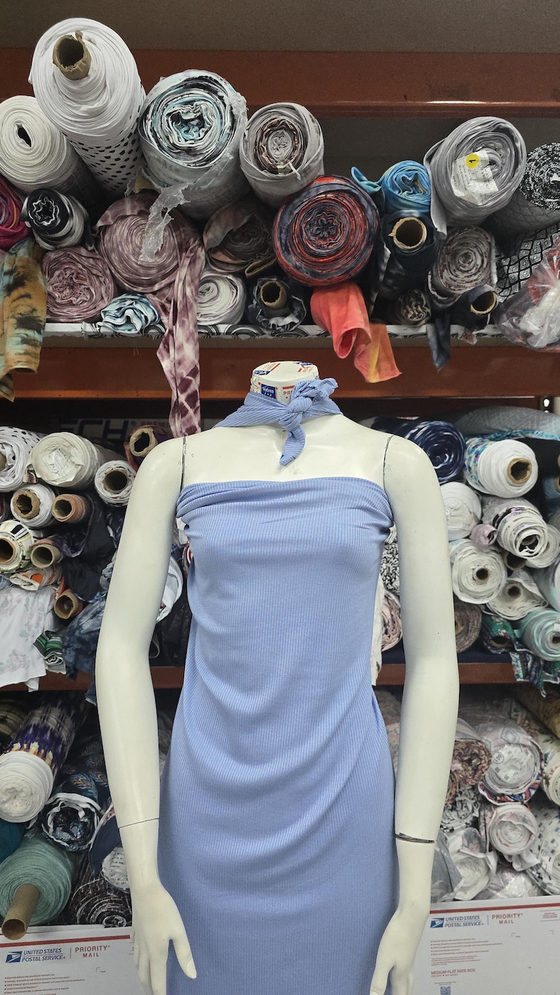 Puede incluir: Un vestido tubo de color azul claro acanalado sobre un maniqu&iacute;, con un pa&ntilde;uelo de cuello a juego de tela acanalada azul claro anudado en la parte delantera. El maniqu&iacute; est&aacute; colocado frente a estantes apilados con rollos de telas.