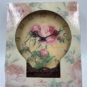 May include: A round Linden wall clock featuring a cream-colored face with a pink rose floral print, black ornate hands, and large dark numerals. The clock is housed in a square cardboard box decorated with matching pink rose patterns and text.