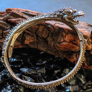 Pu&ograve; includere: Un braccialetto d'argento con un designo dettagliato di testa di drago. Il braccialetto &egrave; aperto e ha una chiusura a cerniera. Il braccialetto &egrave; su una superficie di legno marrone scuro.