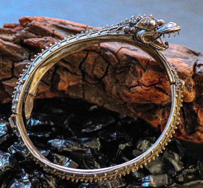 Pu&ograve; includere: Un braccialetto d'argento con un designo dettagliato di testa di drago. Il braccialetto &egrave; aperto e ha una chiusura a cerniera. Il braccialetto &egrave; su una superficie di legno marrone scuro.