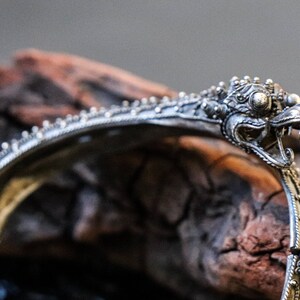 Pu&ograve; includere: Un braccialetto d'argento con un designo dettagliato di testa di drago. Il braccialetto &egrave; ornato e presenta dettagli intricati.
