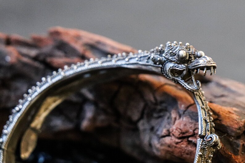 Pu&ograve; includere: Un braccialetto d'argento con un designo dettagliato di testa di drago. Il braccialetto &egrave; ornato e presenta dettagli intricati.