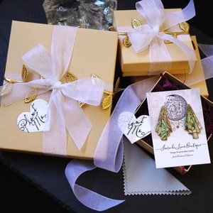 Pu&ograve; includere: Due scatole regalo dorate con nastro bianco e dettagli in foglia d'oro. Le scatole sono legate con un nastro bianco e hanno un'etichetta a forma di cuore che dice "Piantami". La scatola pi&ugrave; piccola ha un'etichetta che dice "Piantami" ed &egrave; appoggiata su una scatola pi&ugrave; grande. Le scatole sono su una superficie nera.