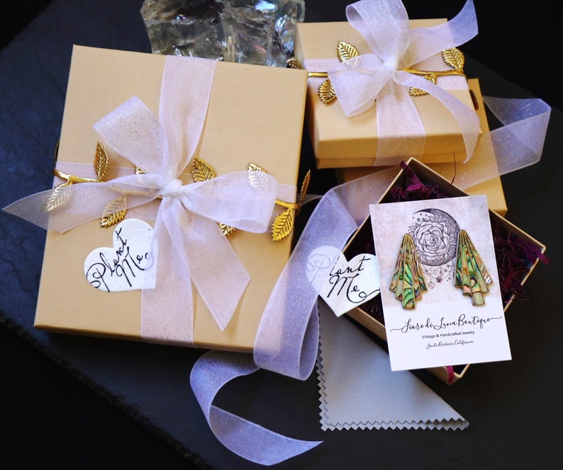 Pu&ograve; includere: Due scatole regalo dorate con nastro bianco e dettagli in foglia d'oro. Le scatole sono legate con un nastro bianco e hanno un'etichetta a forma di cuore che dice "Piantami". La scatola pi&ugrave; piccola ha un'etichetta che dice "Piantami" ed &egrave; appoggiata su una scatola pi&ugrave; grande. Le scatole sono su una superficie nera.