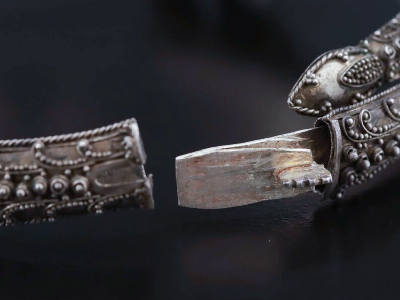 Pu&ograve; includere: Un braccialetto d'argento con disegni intricati e una chiusura a cerniera. Il braccialetto &egrave; aperto, rivelando il meccanismo interno della chiusura.