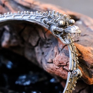Pu&ograve; includere: Braccialetto d'argento con un designo dettagliato e ornato che presenta una testa di creatura mitologica con un becco e denti affilati. Il braccialetto &egrave; aperto e poggia su un pezzo di legno invecchiato.